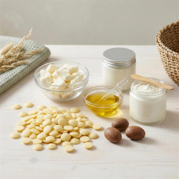 Body Butter Kern Set in Arbeit: geöffneter Kakaobutter-Beutel mit ausgeschütteten Pastillen, offene Sheabutter-Dose, Mandelöl mit Pipette und die fertige Körperbutter in zwei eigenen Gläsern