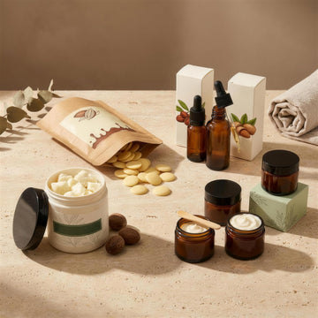 Premium Body Care Set in Arbeit: geöffneter Kakaobutter-Beutel, offene Sheabutter-Dose, Jojobaöl und Mandelöl mit Pipette und vier braune Glastiegel mit fertiger Körperbutter